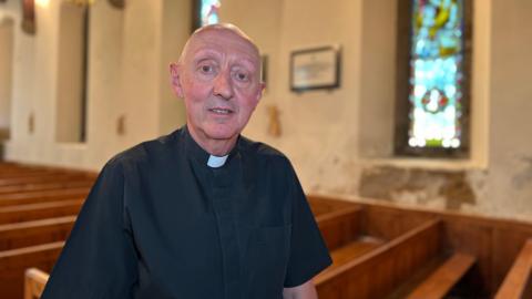 Father Barry looks at the camera in front of a row of pews but in the background there is plaster falling off the wall under a window