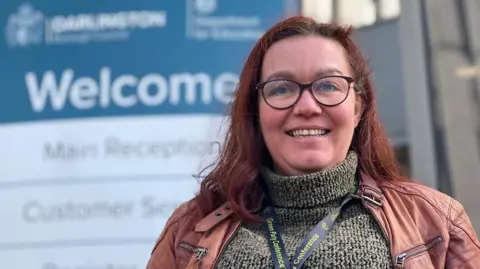 Head and shoulder shot of a smiling Anna-Maria Toms, who has long, dark red hair. She is wearing glasses, a grey polo neck jumper and a brown leather jacket. Round her neck is a  conference lanyard. Behind her, and out-of-focus, is a Darlington Council Welcome sign.