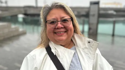 Becky is outside with Belfast Quay behind her. It's been raining, so she's wearing a white raincoat. She has small, gold hoop earrings on and wears glasses. Her shoulder length hair is fair. 