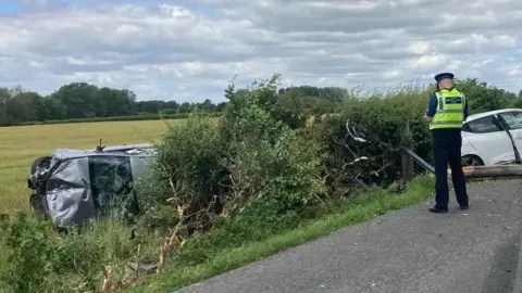 Mike Daniel A crash site on a country road. A silver car has rolled over and is crumpled in a field, by a hedge. There is a police officer and a white damaged car on the right.