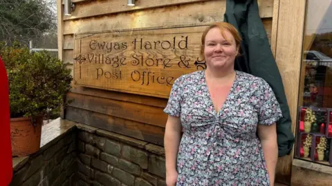 A woman in a blue and pink floral top, standing outside a village shop. There is a large wooden sign to the left that reads "Ewyas Harold Village Shop and Post Office". The woman has short ginger hair and is smiling. 