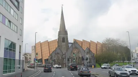 Google A derelict church on a busy roundabout in a city centre. Cars are travelling on the road and roundabout around the church. It is located in front of a large shopping mall. Green traffic lights are on the road around the church.