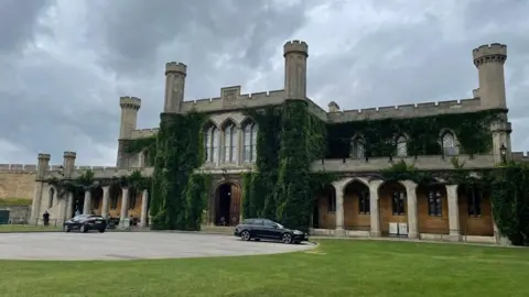 Exterior of Lincoln Crown Court. It's a two-storey Victorian Gothic building with crenellated towers and arches and foliage growing up the walls. Out front is a grass lawn and a roadway with two cars parked on it.