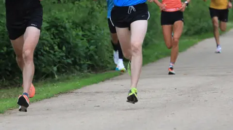 Runners' legs as they pace on a hard path, bordered by grass and hedging.