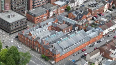 Swindon Borough Council Aerial image of a large building with red brick sides and grey top. Parked cars and a road outside can be seen.