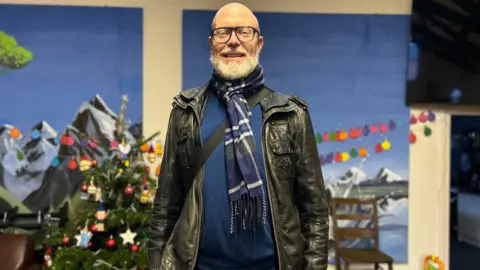 A man stands in front of a wall with Christmas decorations and pictures of mountain scenery. He is bald, wearing glasses, a blue and white scarf, a blue jumper and black leather jacket. The strap of a satchel can be seen going across his body