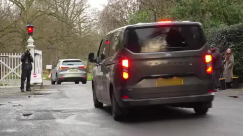 A large grey van follows a smaller silver car through a white gate at Royal Lodge