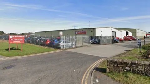 A general view of the  Pollock (Scotrans) yard in Bathgate, West Lothian. It is a sunny day. There are several warehouses in grey with red vehicles parked in front of them. A number of vehicles are parked in a car park at the front of the yard, surrounded by a grey fence.