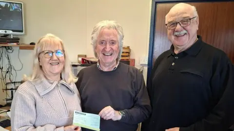 Ronnie Clucas Gillian Jopson stands on the left, from the Queen's Pier Restoration Trust. She had shoulder length grey hair, is wearing a light coloured jumper with a zip into a large collar. and is smiling holding one side of a cheque. Next to her is Roger Corkill on the right holding the other side of the cheque, from the Alan Chinery Trust. He's wearing a dark jumper, is also smiling, with grey hair. On the far right is Queen's Pier Restoration Trust member Graham Curphey, wearing a black jumper, wearing glasses and smiling. He has grey hair at the sides and a grey moustache.