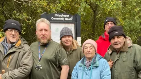 Christine Westerback A group of people in their middle-ages and older stand in front of a board which says Choppington Community Woods. 