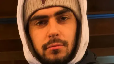 Man looking at the camera he is wearing a woolly hat and a hooded top. He has a short brown beard and moustache.