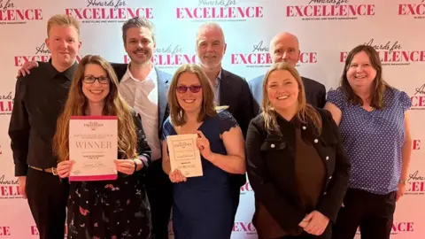 North Somerset Council North Somerset Council and North Somerset Environment Company (NSEC) staff in front of a backdrop saying 'awards for excellence'.
There appear to be four men on the back row wearing shirts and jackets, and smiling towards the camera.
To the right and front row are four women with two wearing dark dresses and two wearing dark blouses and trousers, all smiling at the camera. Two of the women are holding certificates. 