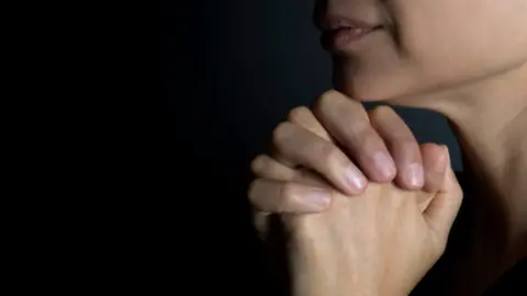 Two hands clasped together in prayer. The mouth and chin of a woman is visible. The rest of the background is dark.