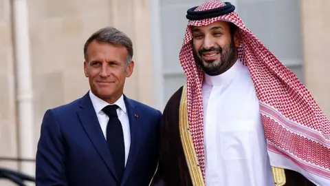 Getty Images A smiling Macron, wearing a navy suit and black tie, poses next to bin Salman, who is wearing traditional Saudi attire