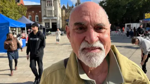 JOHN DEVINE/BBC Toby Wood is looking at the camera. He is wearing a yellow jacket. Behind him is Cathedral Square in Peterborough, with various people walking around temporary market stalls. 
