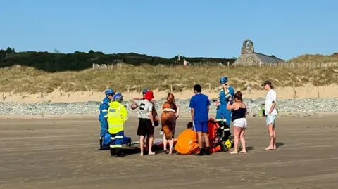 Photograph shows the back of several family members standing onshore at Llandanwg beach. Paramedics can be seen stood among them in brightly coloured blue and white uniforms. 