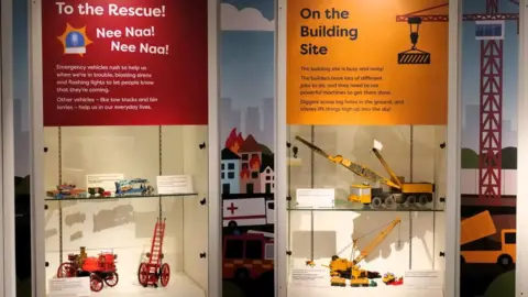 Discovery Museum Two display cabinets, part of a museum exhibition. The one on the left has a red label stating "To the Rescue" and the two shelves below containing models including a Victorian fire engine and an extendable ladder on wheels. The one on the right has a yellow label stating "On the Building Site" and its models include a lorry with lifting gear and a crane on caterpillar tracks.