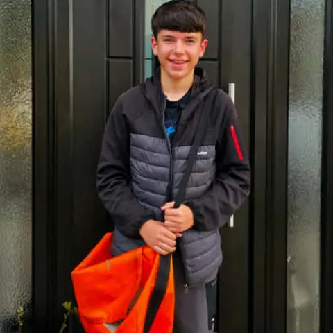 Callum Clydesdale, a dark-haired young boy, stands outside his front door - a black modern door with glass in the centre. He is wearing a grey and black anorak and his large papers delivery bag, which is bright orange.