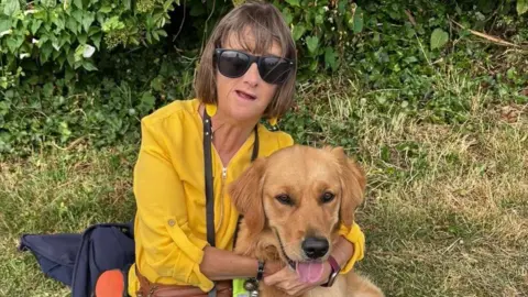 A woman in a yellow top cuddles a dog which has its tongue out