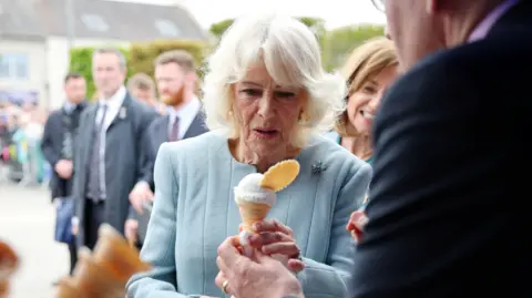Reuters Britain's Queen Camilla meets local traders during her visit to Conway Square, in Newtownards. She is wearing a light blue jacket with brooch. She is being handed an ice cream cone with wafer.