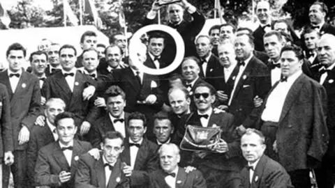 Llangollen International Eisteddfod A black and white image of a group of about 40 Italian choristors from Corale Rossini. The men all wear suits and bow ties. The front rows are crouched down. At the centre of the photo is a young Luciano Pavarotti. A white circle has been drawn around him to highlight who he is. He is smiling straight at the camera and has short dark hair.