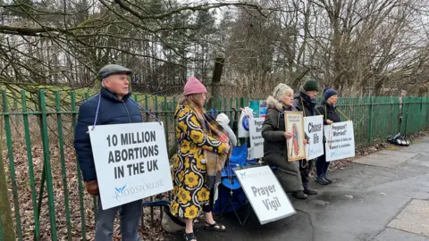 Five people standing against a park fence outdoors, all holding signs saying things like 10 MILLION ABORTIONS IN THE UK and PRAYER VIGIL