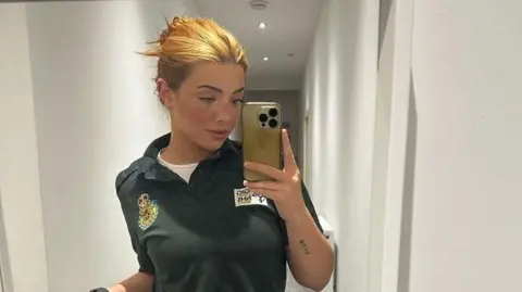 Lorna is using her mobile phone to take a selfie in a mirror here. She has blonde/ginger hair which is pinned up. She's wearing a green Welsh ambulance service polo shirt with a white shirt underneath. 