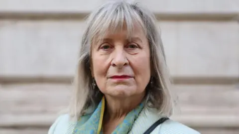A woman with shoulder length greying hair and a pale green jacket stares directly at the camera