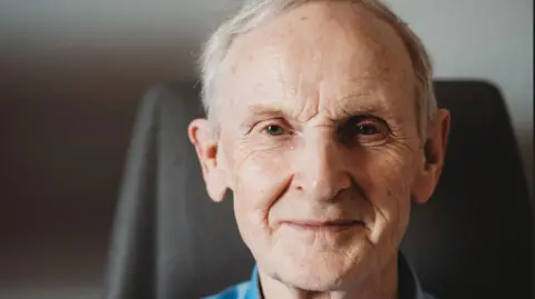 Headshot of an elderly man, with white hair and wearing a blue shirt and standing in front of a fireplace in a sitting room