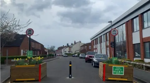 A couple of wooden plantgers, full of daffodils, with a bollard in the middle preventing traffic from getting down a terraced road.