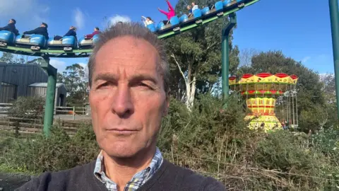 Rick Turner A man stands in front of a rollercoaster at a visitor attraction. You can see people on the amusement ride with their hands in the air. There is also a carousel in the background.