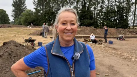 Melanie Stanford standing to one side of an excavated area. She is wearing a blue body warmer and t-shirt and is smiling at the camera.