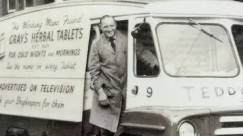 BBC John Gray stands in the door of an old-fashioned van with The working man's friend Gray's Herbal Tablets for Cold Nights and Mornings written on the side. Gray is smiling and wearing a long apron, shirt and tie. 