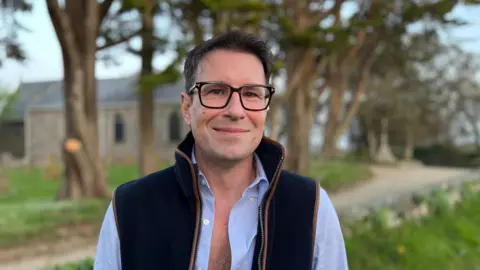 Christopher Kennedy-Barnard - A man with black hair, square rimmed glasses wearing a blue and brown rimmed gillet, on top of a blue button up shirt where many of the buttons are undone. 
