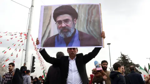 EPA An Iranian man, in a black suit jacket and white shirt, holds a picture of Mojtaba Khamenei, as he takes part in celebrations of the 47th anniversary of the Islamic Revolution in Tehran on 11 February 2026.
