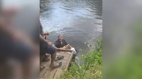 Samer Jouha holding onto an elderly woman in the River Avon in Evesham. He is  holding onto her as he looks up at a short wooden jetty, where other people are coming to assist him.
