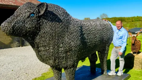 BBC Steven Nesbitt standing at his farm with his arm around his big metal bull. He is wearing a light blue shirt and grey jeans, and has a shaved head. It is a bright sunny day.