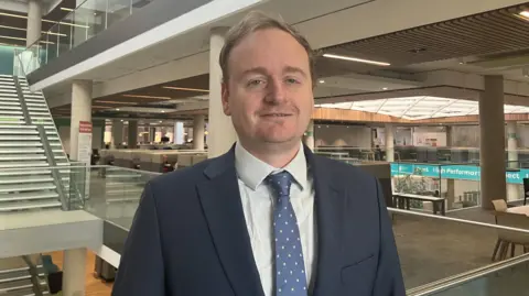 Nadia Lincoln/ LDRS Adam Brown, in a navy suit, a navy tie with yellow spots on, and a pale blue shirt, stands in the council's offices.