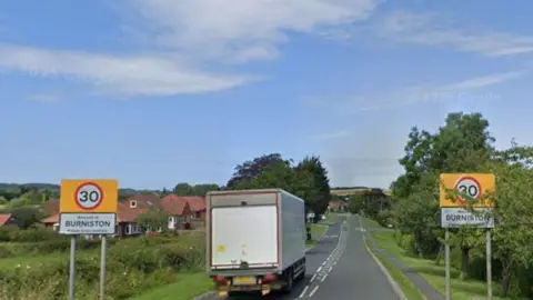 Google Two sign posts displaying town name of Burniston and a 30mph speed limit with a lorry driving between them