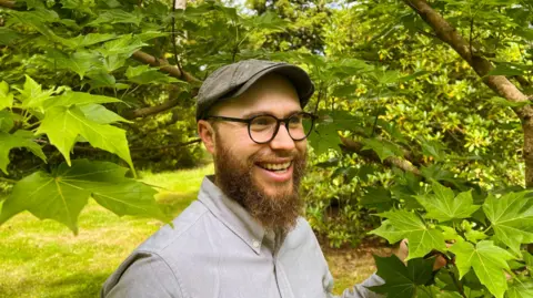 Dan Crowley Dan Crowley stands among branches and leaves. He wears a flat cap, dark-rimmed glasses and a brown beard