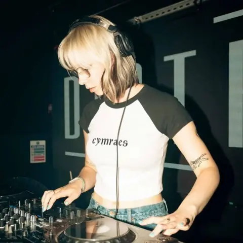Molly Palmer A woman with short blonde hair and glasses, looking down at DJ decks. She is wearing a white and black t shirt which reads: Cymraes.