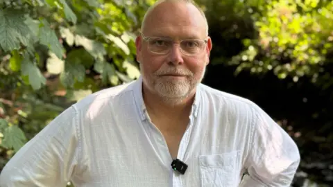 Gerry Darby has a short white beard and short white hair at the sides of his head. He is wearing glasses with a see through frame and a white shirt which is unbuttoned at the collar. He is standing outside in front of some trees.