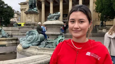 Yume Penwarden is pictured standing in front of the Victoria Rooms in Bristol. She is a young woman with dark hair that is tied back. She is wearing a bright red T-shirt underneath a red University of Bristol hoodie. She is smiling.

