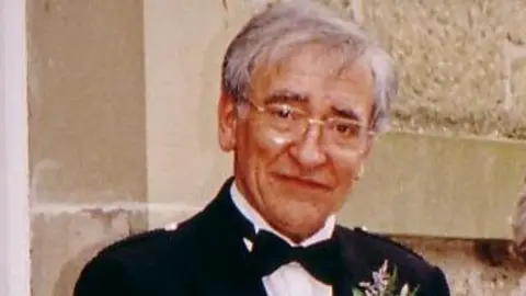 A man in a dark jacket, white shirt and bow tie standing in front of a wall