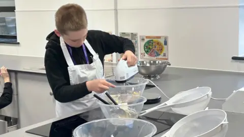 Kate Bradbrook/BBC Tyler, using a whisk, in a school kitchen, with a number of clear bowls in front of him, he is looking down. There are two sieves on the counter and a silver bowl behind him. He is wearing a black jumper, with a white apron over the top. 