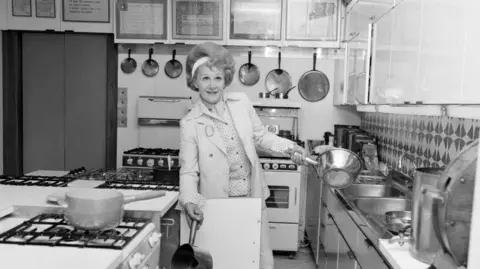 Getty Images A well dressed woman holding two saucepans in a large kitchen surrounded by gas stoves and sinks with frying pans hanging on the wall in the background