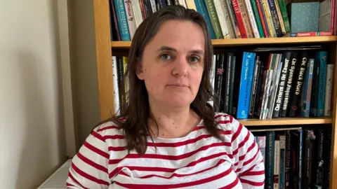 BBC A woman with long brown hair is wearing a white and red striped t-shirt. She is standing in front of a full bookshelf and is next to a cream wall and radiator.