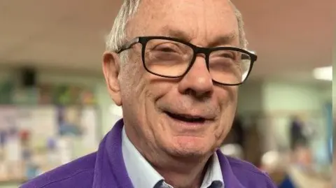 A close-up of Tony smiling at camera, wearing a purple fleece with the BDAA logo on it and with the memory cafe taking place behind in a blurred background