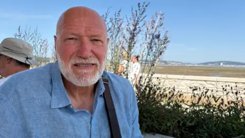 A man in a blue shirt with a white beard sat on Mumbles seafront. 