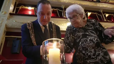 A man in a dark suit stands indoors wearing an ornate gold ceremonial chains, standing next to an elderly woman with white hair. She is wearing a black top. They are lighting a candle.